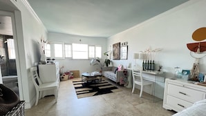 Amplia sala de estar con sofá, una mesa de centro y un comedor con una mesa y sillas blancas. Se observan ventanas que permiten la entrada de luz natural, y una planta en la habitación.