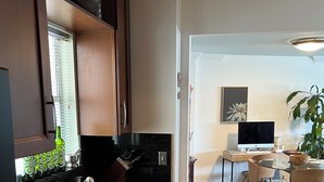 Una cocina moderna con armarios de madera oscura, encimera de granito negro y pileta de acero inoxidable. Hay una zona de comedor con mesa y sillas, y una planta grande en maceta.