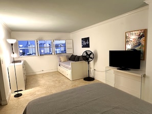 Un dormitorio con una cama, televisor, ventilador, cómoda y ventana con vista a la ciudad.