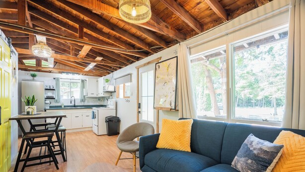 Una cocina moderna con vigas de madera, un comedor con mesa y sillas, un sofá con cojines y ventanas amplias con vistas a árboles.