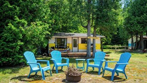Una cabaña de color amarillo con un porche, rodeada de árboles. Cuatro sillas Adirondack azules están dispuestas alrededor de un brasero.