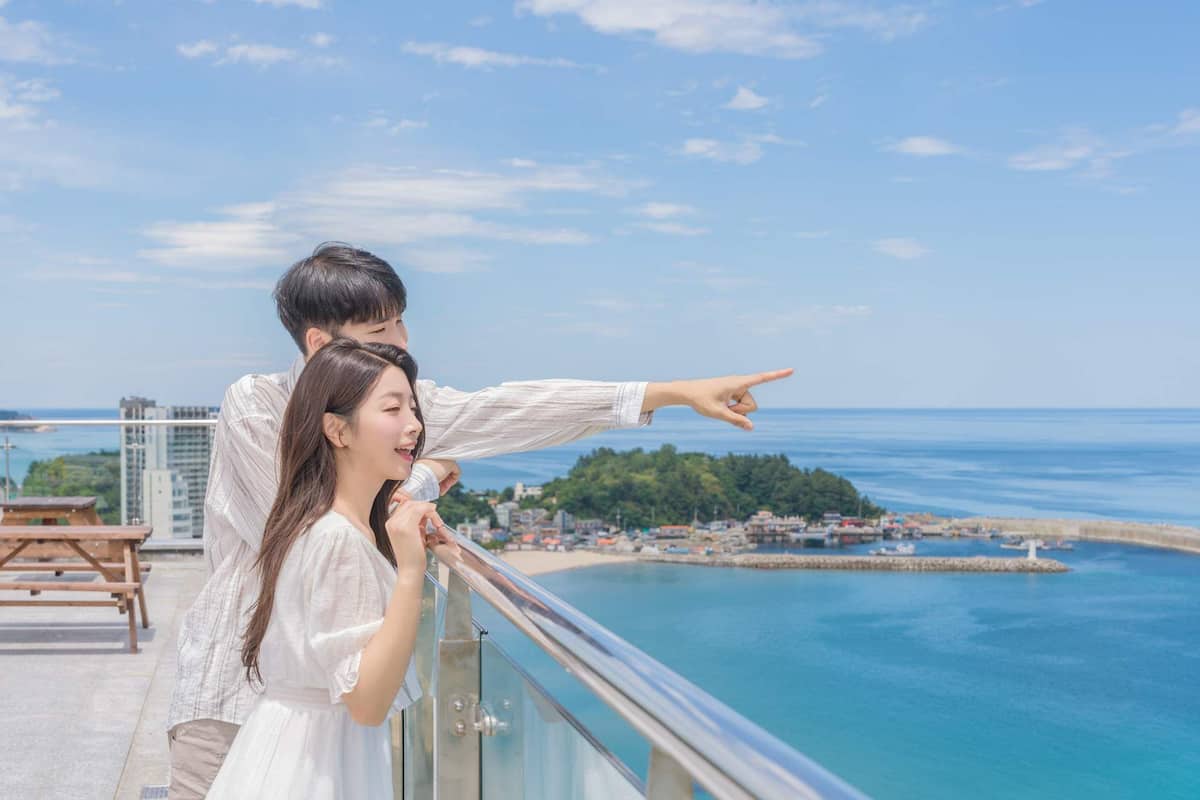 A couple on a balcony overlooking a coastal view with buildings and a beach in the distance.