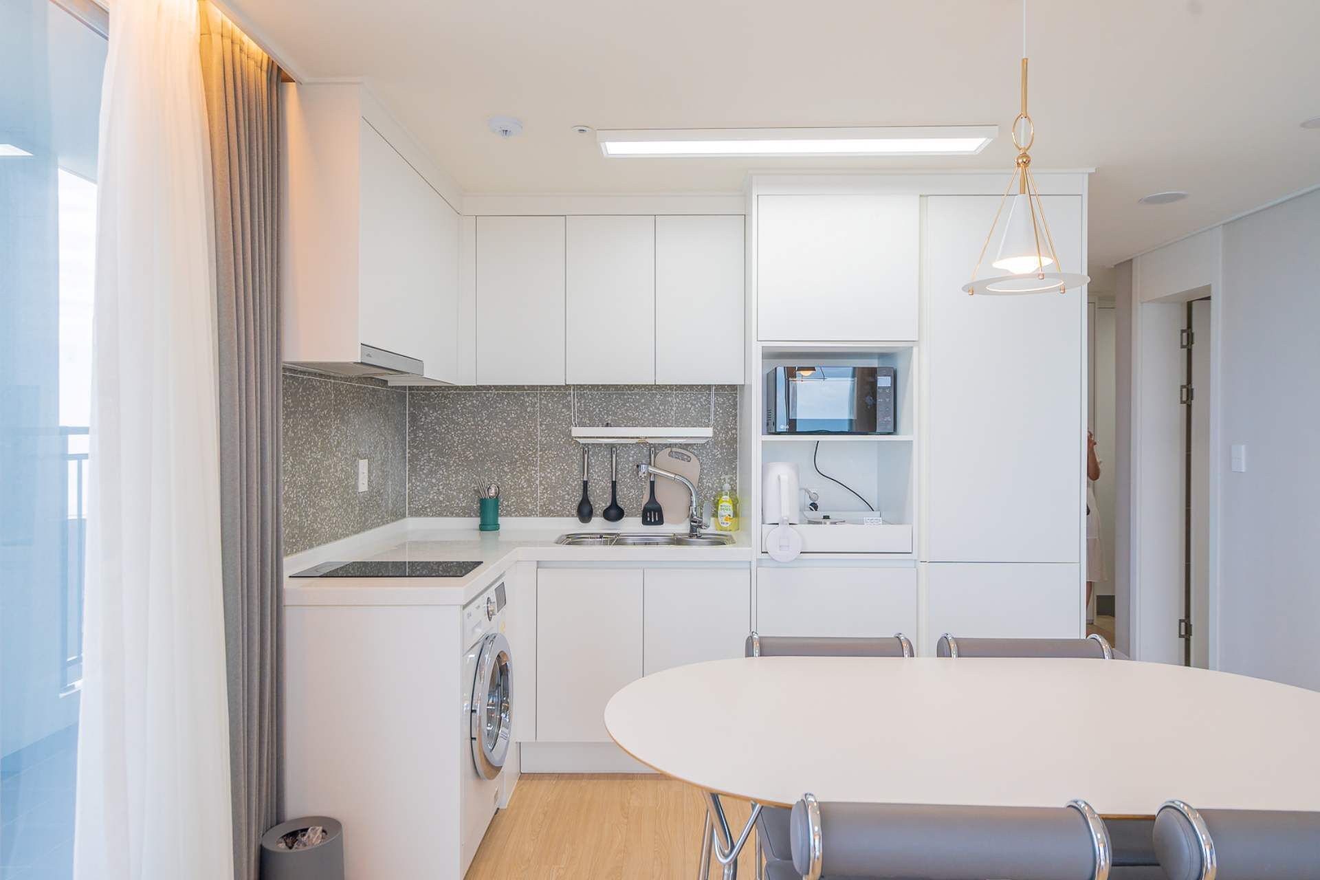 A modern kitchen with white cabinets, a built-in oven, a microwave, and a sink. There is a dining table with chairs and a hanging light fixture.
