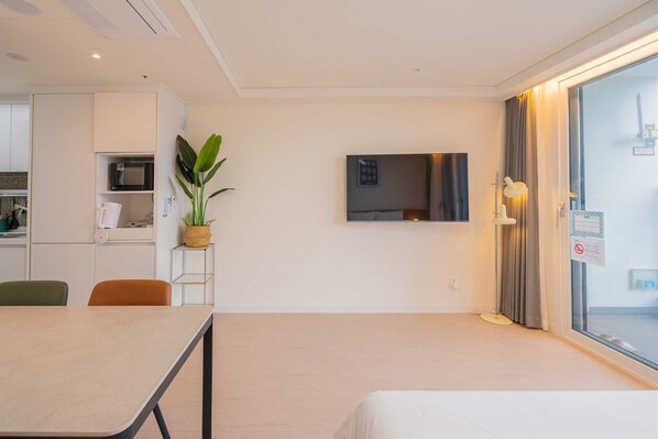 Una habitación de hotel moderna con zona de comedor, televisor y un amplio ventanal con cortinas.