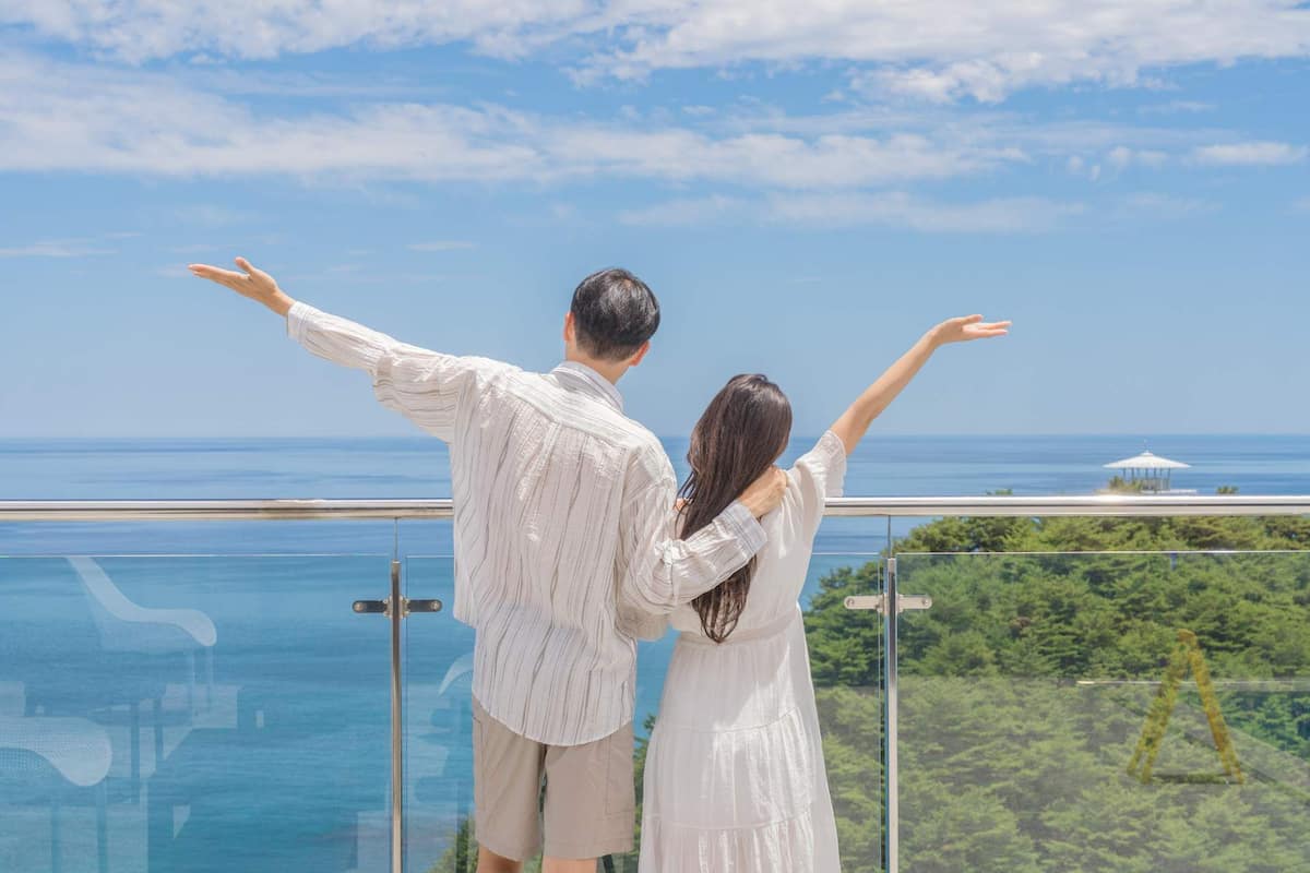 A couple standing on a balcony with a clear view of the ocean and greenery below.