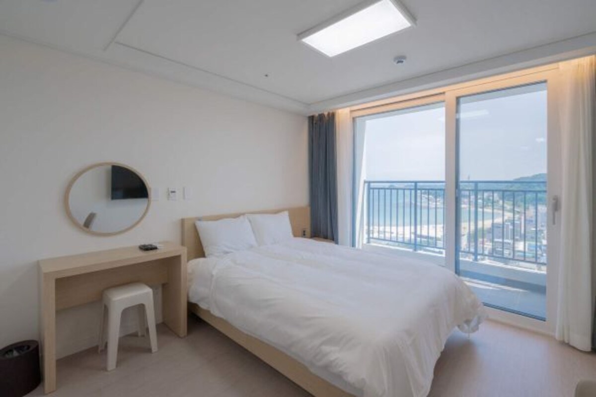 Habitación de hotel con cama, escritorio, silla, espejo y ventana grande con vistas al exterior.