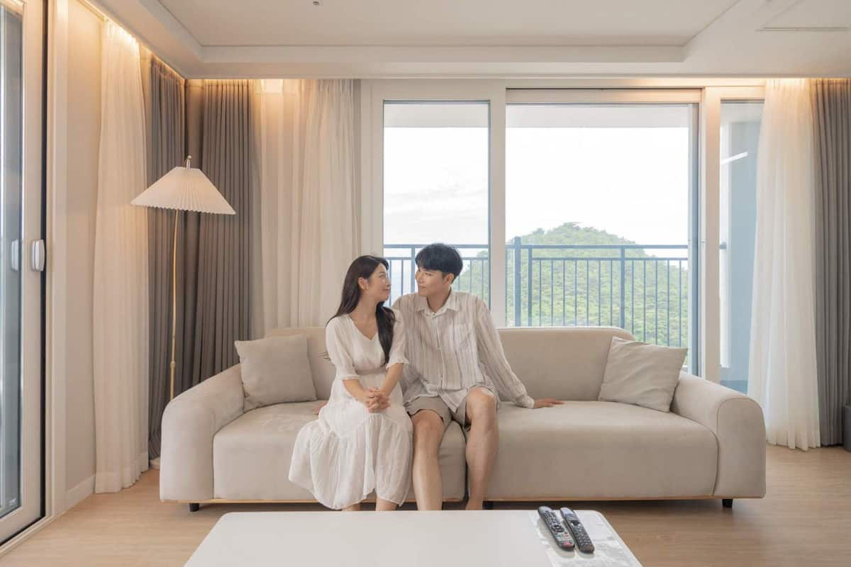 A couple sitting on a sofa in a well-lit room with large windows and curtains.
