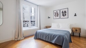 A bedroom with a large window, a bed with a blue blanket, two bedside tables, and two wall-mounted lamps.