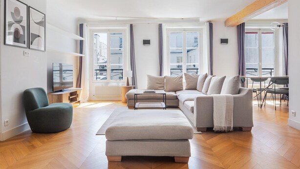 A modern living room with a sofa, ottoman, and a green chair. There are large windows with curtains, a radiator, and a TV on a stand.