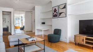 A modern living room with a sofa, a coffee table, a TV stand, and a chair. There is a bookshelf and a door leading to another room.