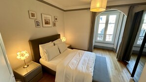 Une chambre à coucher avec un lit, des lampes de chevet, une table de nuit, une fenêtre avec des rideaux et des tableaux encadrés accrochés au mur.