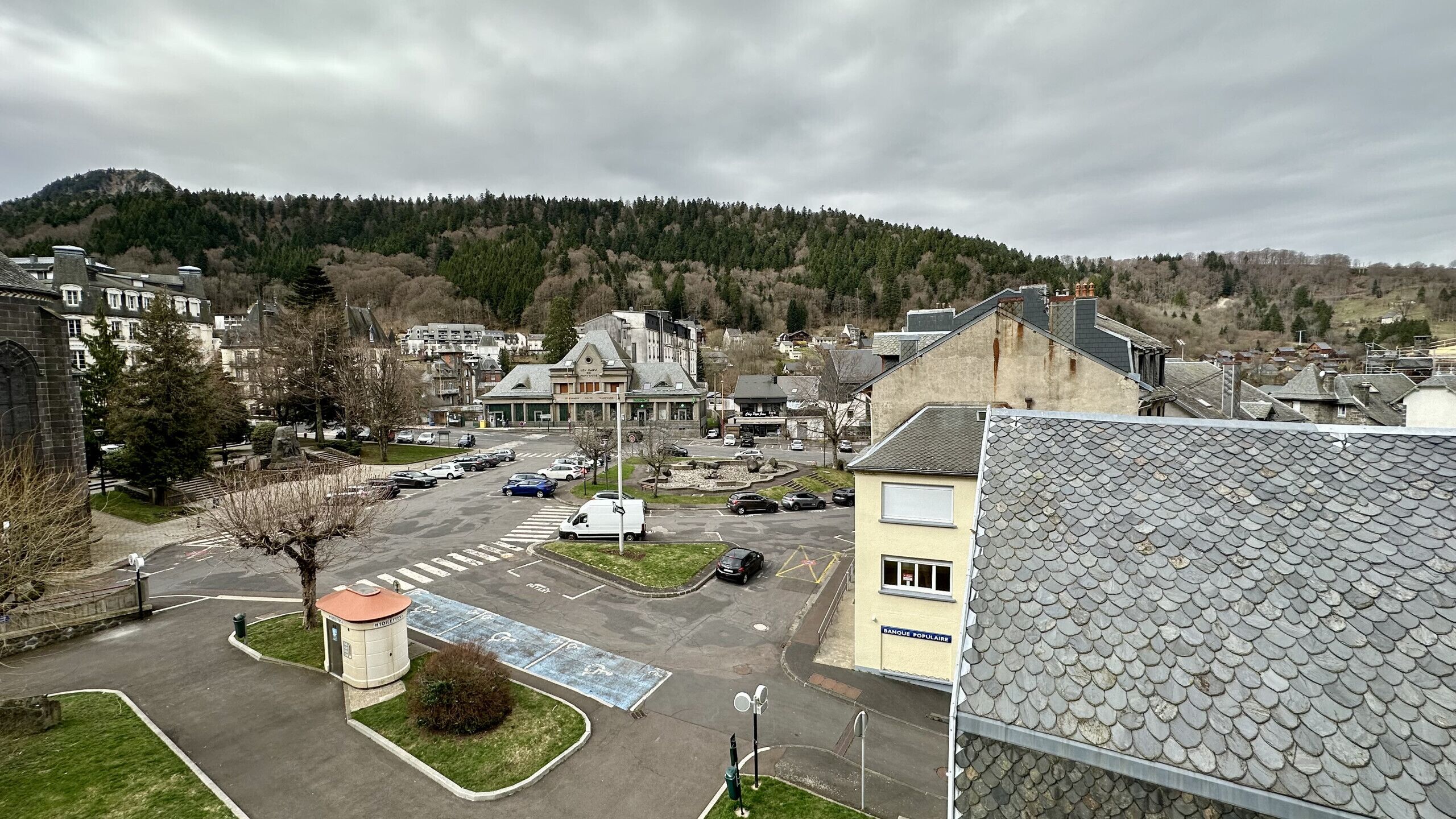 Un parking rempli de voitures, un petit bâtiment avec une enseigne indiquant « BANQUE POPULAIRE », et une colline avec des arbres en arrière-plan.