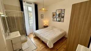 A bedroom with a bed, a bedside table with a lamp, a shower area, and a window with curtains.