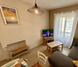 A living room with a television, a wooden table, a bench, a sofa, and a window with curtains. - Villa Michel Le Mont Dore 10pers (Mont-Dore)