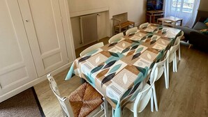 Un coin repas avec une table dressée pour quatre personnes, des chaises blanches, un radiateur, un téléviseur posé sur un meuble et une fenêtre avec des rideaux.