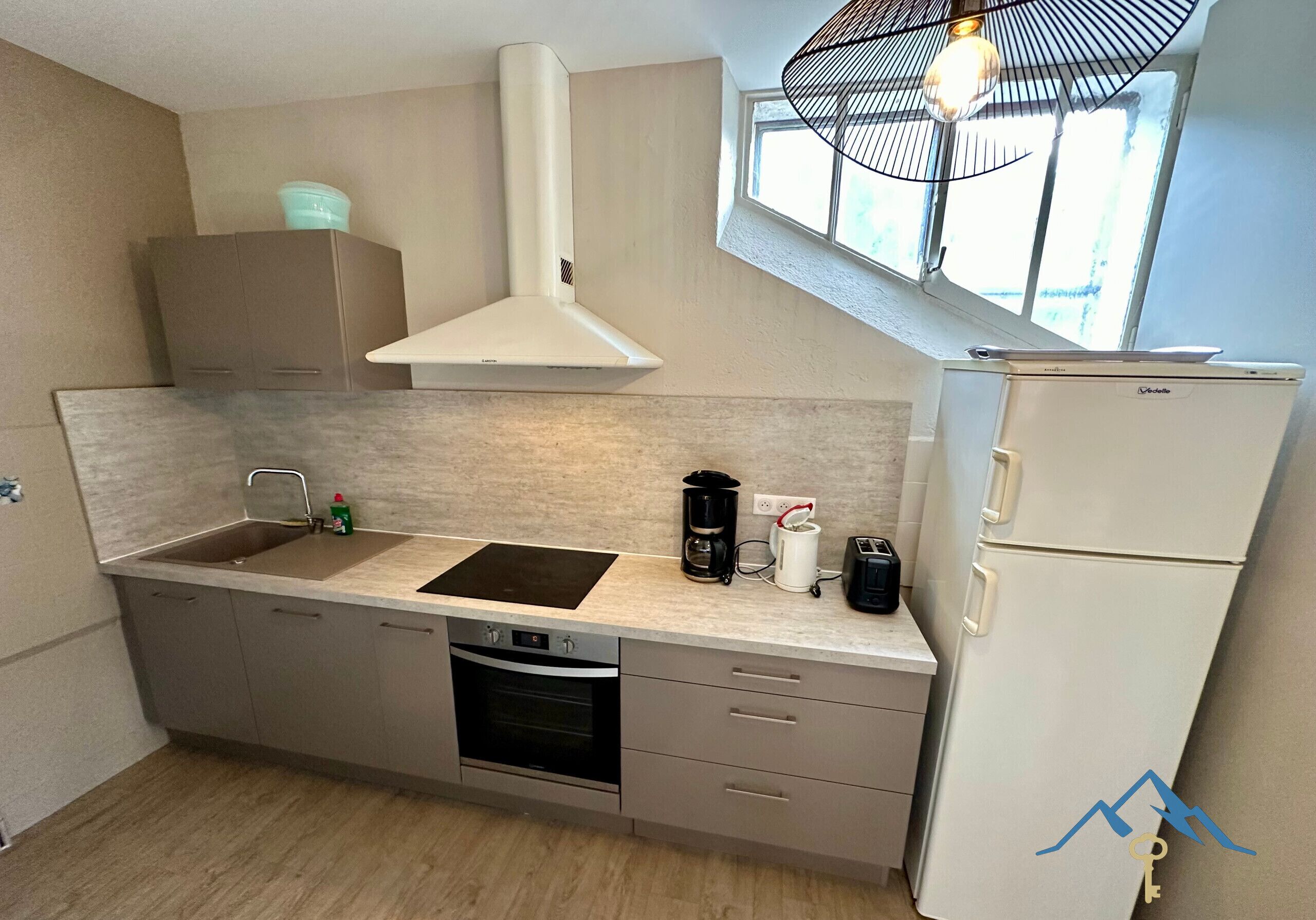 Cocina compacta con lavamanos, estufa y refrigerador, con ventanal sobre el lavamanos para luz natural.