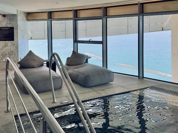 Un área de piscina interior moderna con vistas al mar, un salón con dos sofás grises y una escalera metálica que lleva a la piscina.