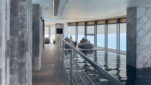 Un área de piscina interior moderna con paredes de mármol, barandilla metálica y vistas al océano.