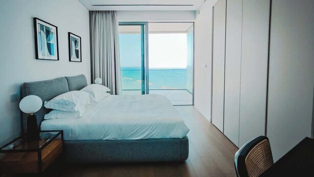 Una habitación de hotel moderna con una cama grande, una mesita de noche, una silla y vista al océano a través de un ventanal.