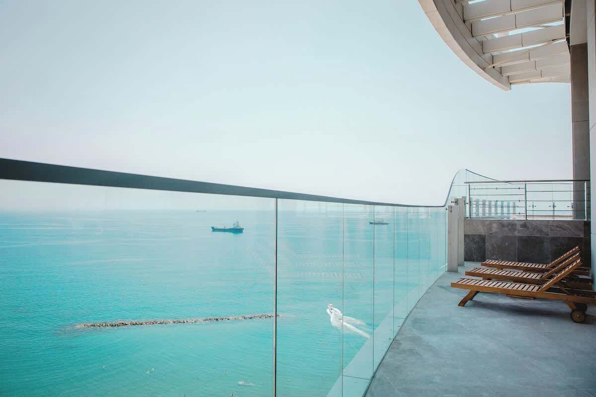 Een balkon met glazen balustrade, uitzicht op een wateroppervlak, een houten lounge stoel en een heldere lucht.