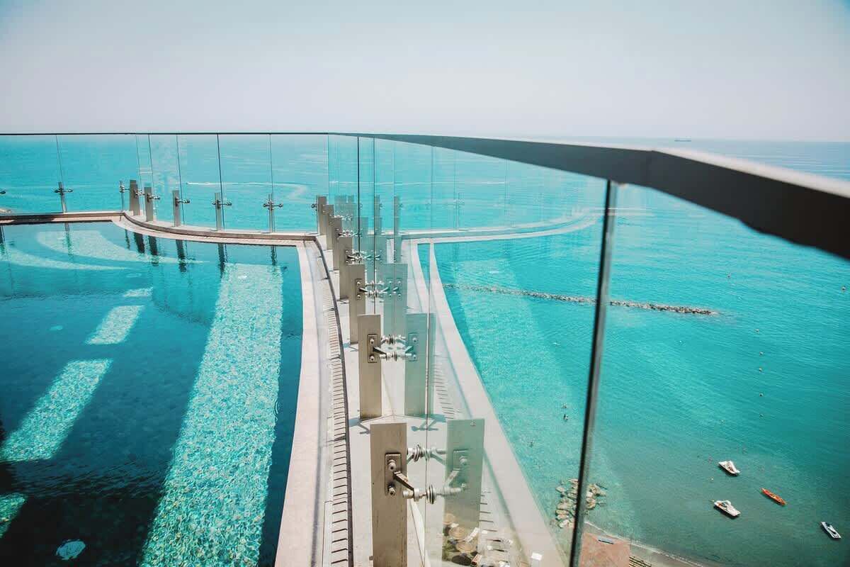 A glass-enclosed infinity pool with clear blue water, a curved deck, and a view of the sea.