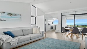 Uma sala de estar moderna com um sofá cinzento, uma área de jantar com mesa e cadeiras, e uma cozinha ao fundo.