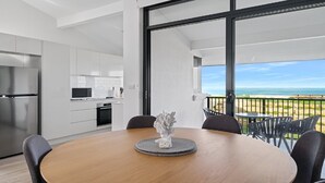 Uma cozinha moderna com armários brancos e eletrodomésticos de aço inoxidável, uma área de refeições com mesa e cadeiras de madeira e uma varanda com vista para a praia.