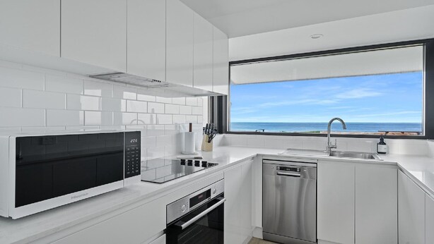 Uma cozinha moderna com armários brancos, micro-ondas embutido, forno e lava-louças, e uma grande janela com vista para o oceano.