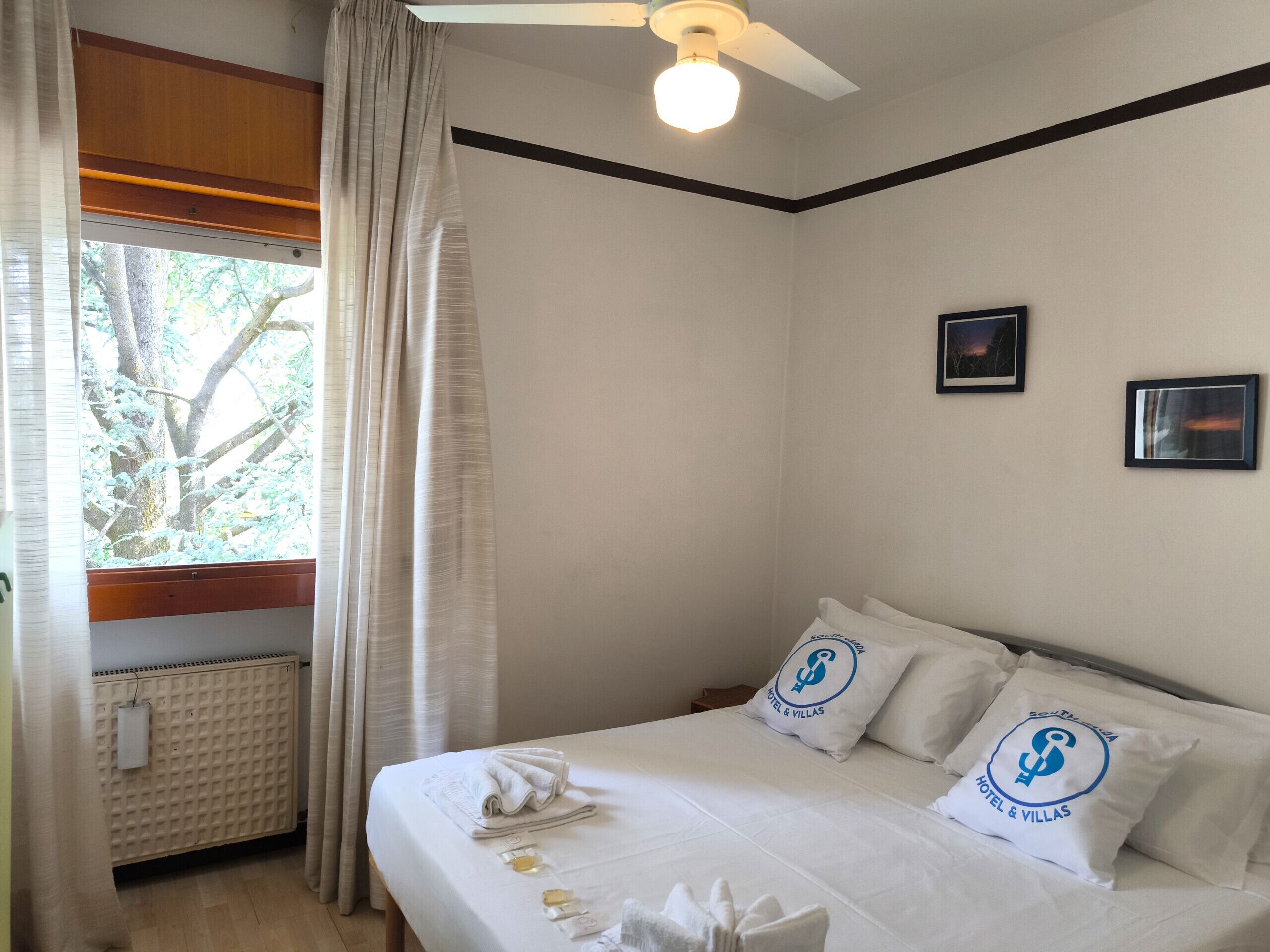 A hotel room with a bed, two pillows with logos, a window with curtains, a ceiling fan, and a radiator.