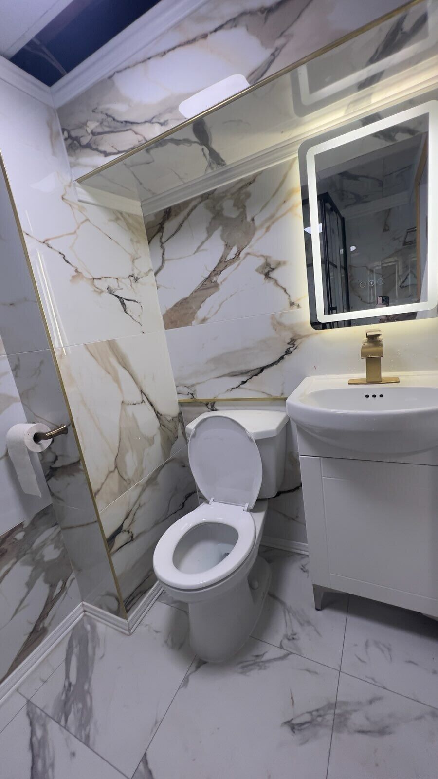 Un baño con piso de mármol, inodoro y lavamanos blancos, y un espejo con luz.