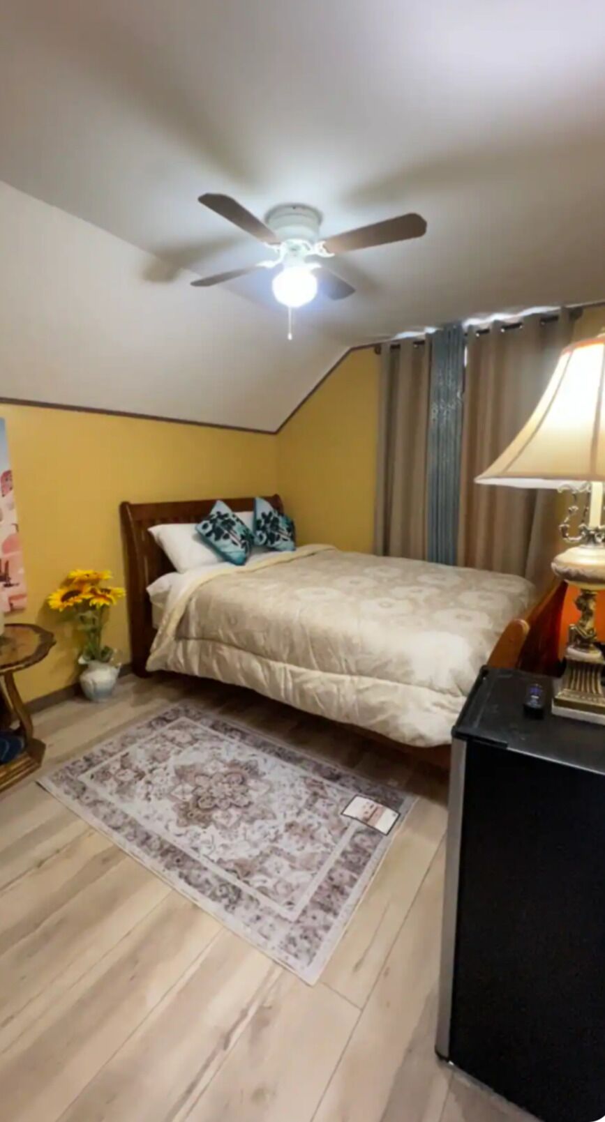 Une chambre à coucher comprenant un lit, un ventilateur de plafond, une table de chevet, une lampe et une petite table sur laquelle est posé un vase de fleurs.