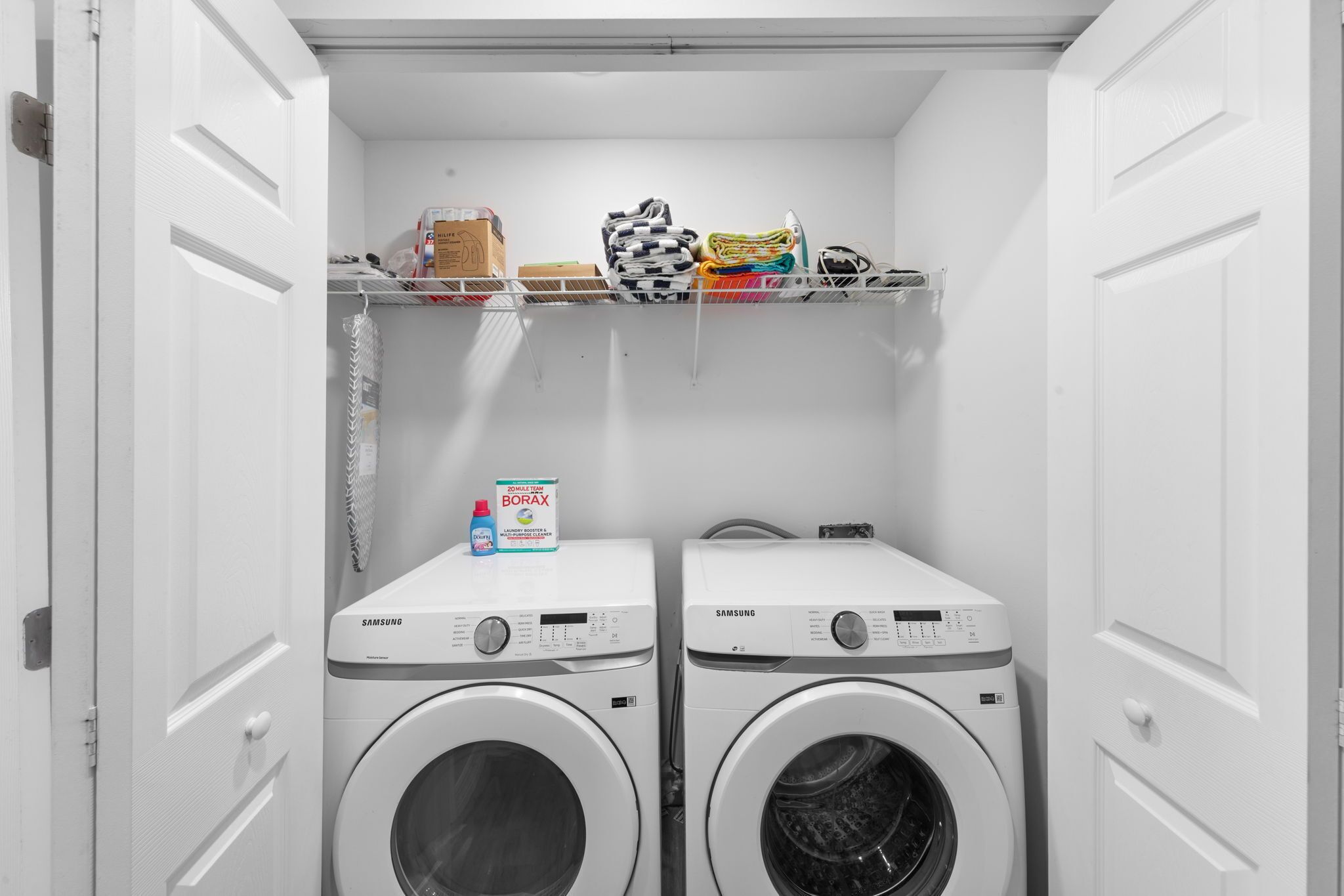 Een kleine wasruimte met twee Samsung wasmachines, een plank met opgevouwen kleding en een doos Borax wasmiddel.
