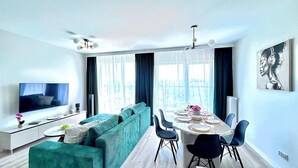 Un salon moderne doté d’un grand téléviseur, d’un canapé turquoise, d’une table à manger pour quatre personnes et offrant une vue sur la ville grâce à de grandes fenêtres.
