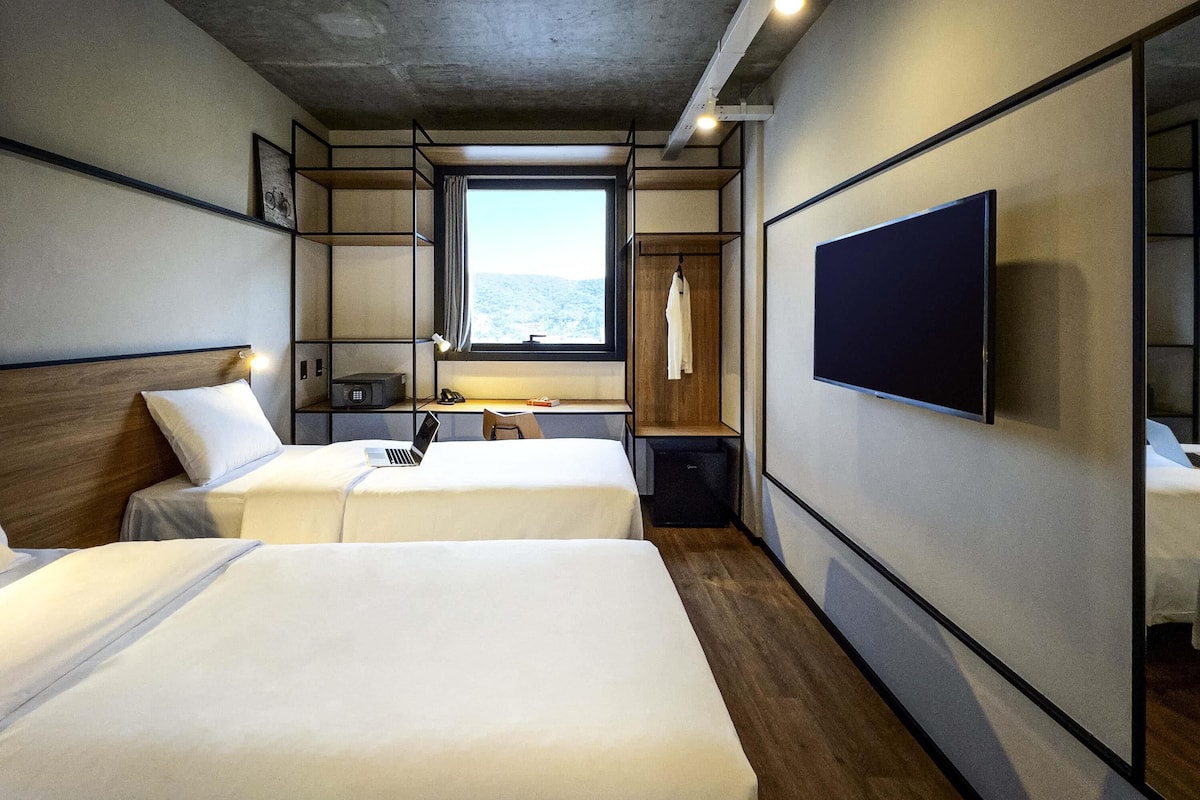 A modern hotel room with two beds, a TV, a desk with a chair, a window with a view, and a wardrobe.