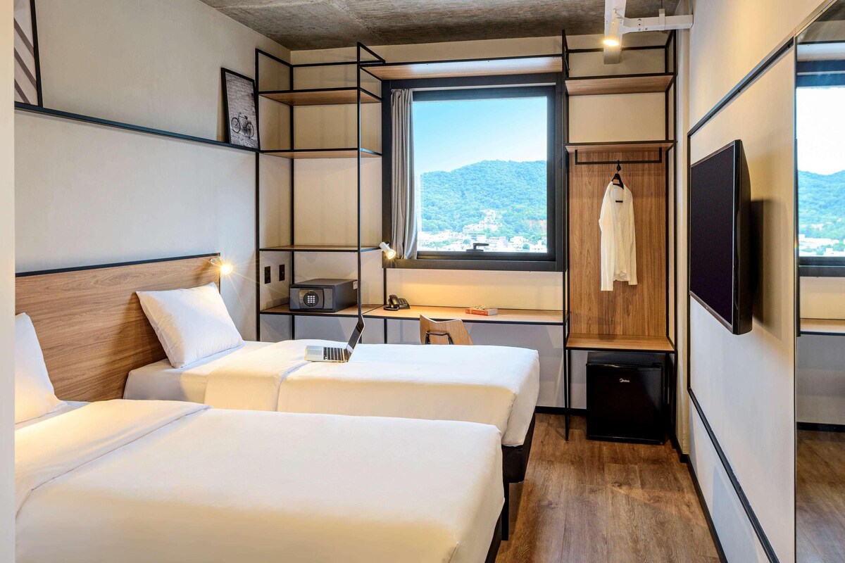 A hotel room with two beds, a desk, a chair, a TV, and a window with a view of the mountains.