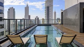 Una piscina en la azotea con tres sillas de descanso, rodeada por una barandilla de cristal, con vistas a un paisaje urbano de edificios altos.