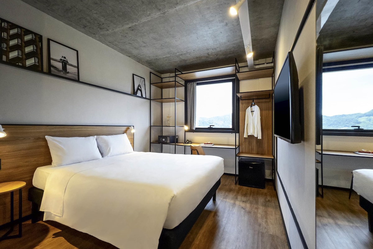 Ein modernes Hotelzimmer mit einem großen Bett, einem Schreibtisch, einem Stuhl, einem Fernseher und einem Fenster mit Blick.