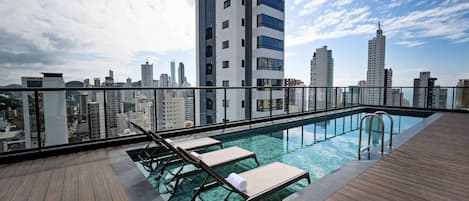 Una piscina en la azotea con cubierta de madera, sillas de descanso y vista al horizonte de la ciudad.