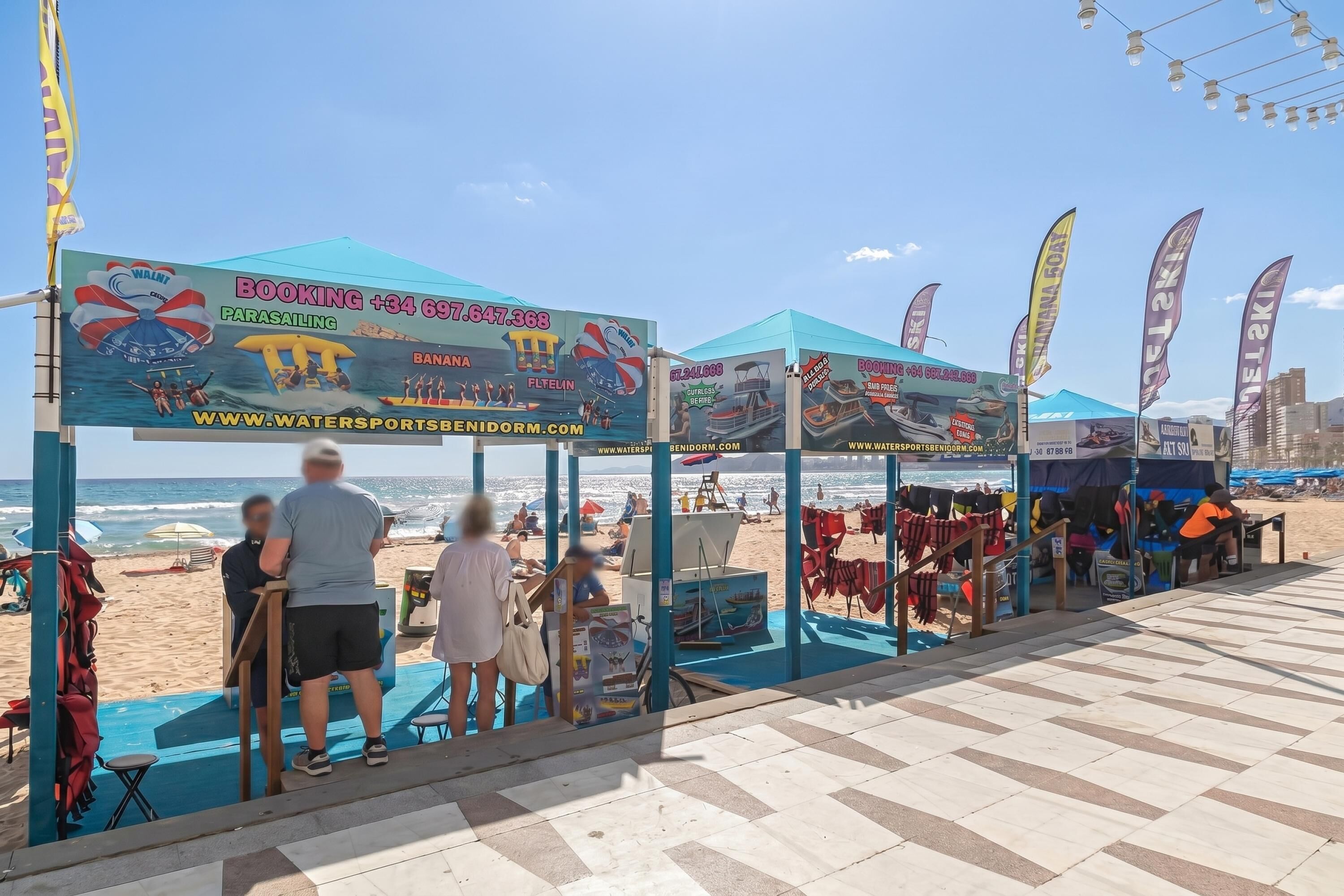 Zona de playa con parapentes, equipo para deportes acuáticos, tiendas de campaña y sillas de playa.
