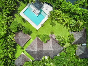 An aerial view of a resort with a swimming pool, thatched-roof structures, and lush greenery.