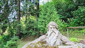 A stone statue of a bearded figure on a moss-covered rock in a forested area.