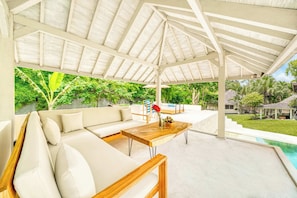 A covered outdoor seating area with a wooden table, a sofa, and a view of a swimming pool and lush greenery.