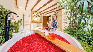 A bathroom with a bathtub filled with rose petals, a wooden shelf with a pitcher of water, and a person reading a book. - Villa Indigo - 5-BDR, huge pool and lush garden (Kecamatan Kuta)