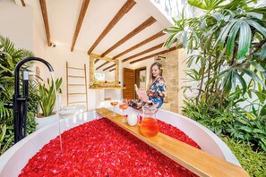 A bathroom with a bathtub filled with rose petals, a wooden shelf with a pitcher of water, and a person reading a book. - Villa Indigo - 5-BDR, huge pool and lush garden (Kecamatan Kuta)