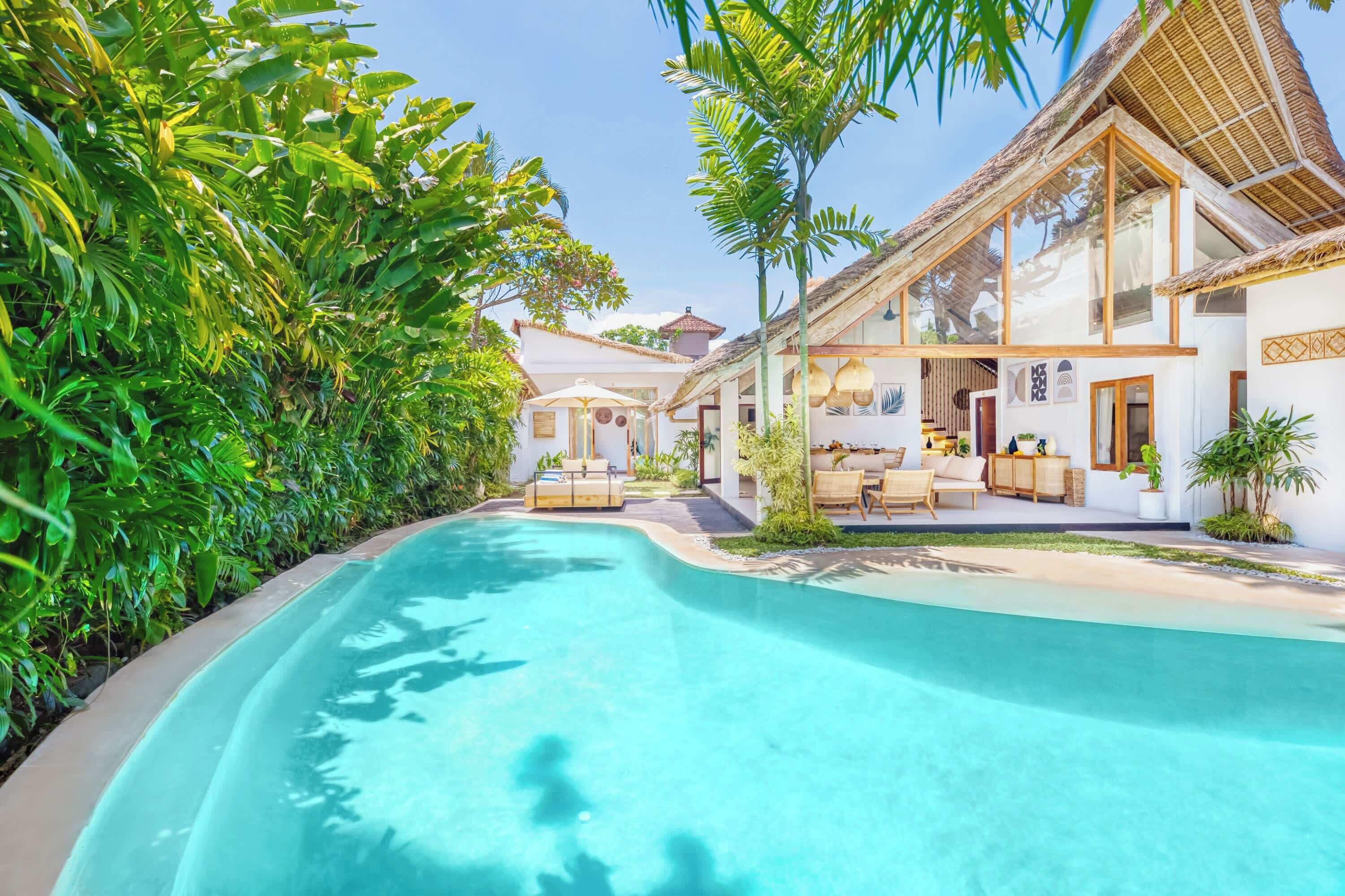 A poolside area with a clear blue pool, surrounded by lush greenery and a building with large windows and outdoor seating.