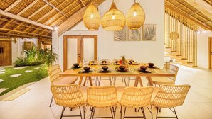 A dining area with a long table set for a meal, surrounded by wicker chairs, under a thatched roof. - Villa Indigo - 5-BDR, huge pool and lush garden (Kecamatan Kuta)