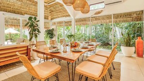 A spacious room with a large dining table set for six, surrounded by wicker chairs, a sofa, and a large potted plant.