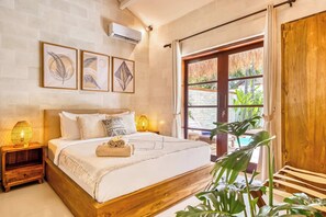 A bedroom with a wooden bed frame, a nightstand with a lamp, framed artwork on the wall, and a view of greenery outside.