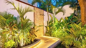 A landscaped garden with a stone wall, various tropical plants, and a paved pathway. - Villa Aurora - 6BDR, near beach, big pool & garden (Kuta)
