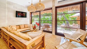 A modern living room with a sofa, a coffee table, wicker chairs, a TV, and large windows with a view of greenery. - Villa Aurora - 6BDR, near beach, big pool & garden (Kuta)